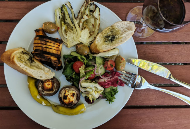 Vegetarisches Barbecue, Teller und Weinglas mit Fenchel, Champignons, Oliven und Kräuterbaguette.